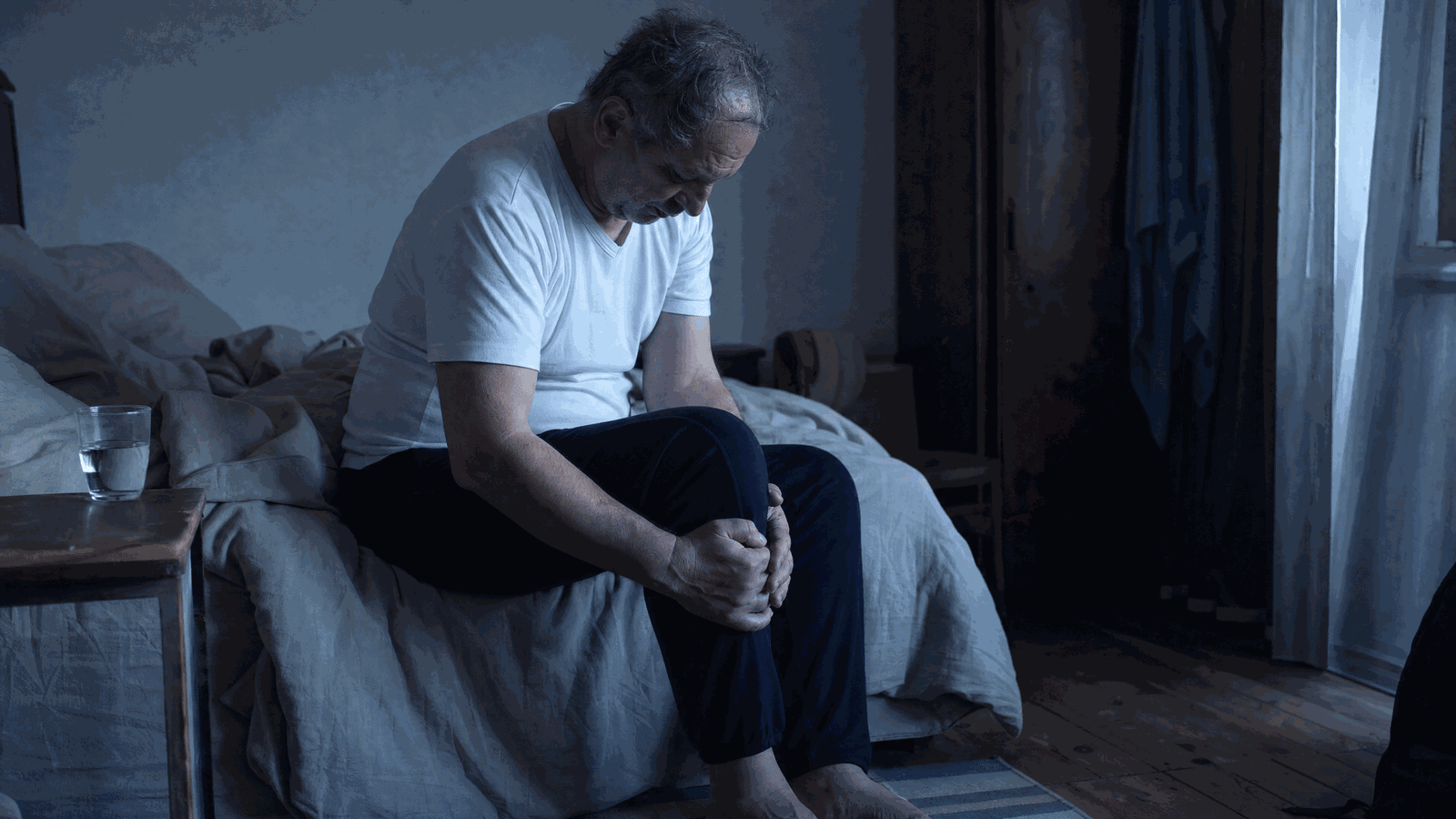Uomo seduto sul bordo del letto all’alba con le mani sul ginocchio rigido